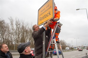 Rostocks Verkehrssenator Holger Matthäus und Oberbürgermeister Roland Methling beobachten, wie Rektor Prof. Schareck dabei hilft, das erste neue Ortseingangsschild mit dem Namenszusatz Hanse- und Universitätsstadt anzubringen.