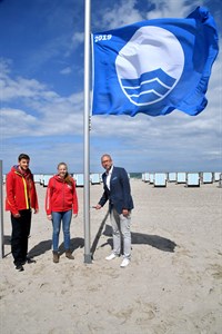 Die DRK-Rettungsschwimmer Max Cornelius und Sarah Wenzel hissen gemeinsam mit Matthias Fromm, Tourismusdirektor von Rostock & Warnemünde, die Blaue Flagge am Strand von Warnemünde (Foto: TZRW/Joachim Kloock)