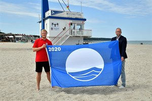 Eike Reck, Rettungsschwimmer der DRK Wasserwacht, und Matthias Fromm, Tourismusdirektor Rostock & Warnemünde, präsentieren die Blaue Flagge 2020 (Foto: TZRW/Joachim Kloock)